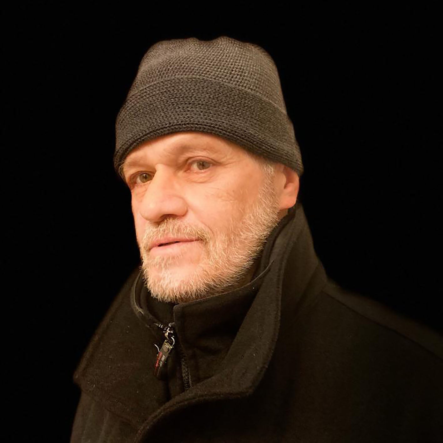 Alter Mann mit weißen Bartstoppeln trägt eine Graue Beanie-Mütze. Aus Merinowolle quer in Kopf-Form gestrickt. Das Fangpatenmuster bedeutet daß Innen ein Knötchenmuster und außen ein klasiches Rechte Maschenmuster zu sehen ist. Das rote vincente-Label mit der kleinen Ente ist am Rand angenäht. 