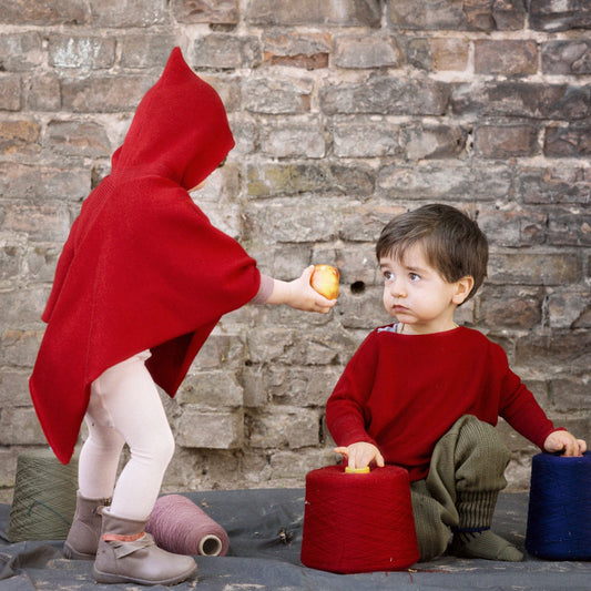 Ein kleines Mädchen mit einem roten Cape mit Kapuze reicht einem kritisch drein blickenden Jungen  einen Apfel. Der Junge spielt lieber weiter mit den Wollkonen.
