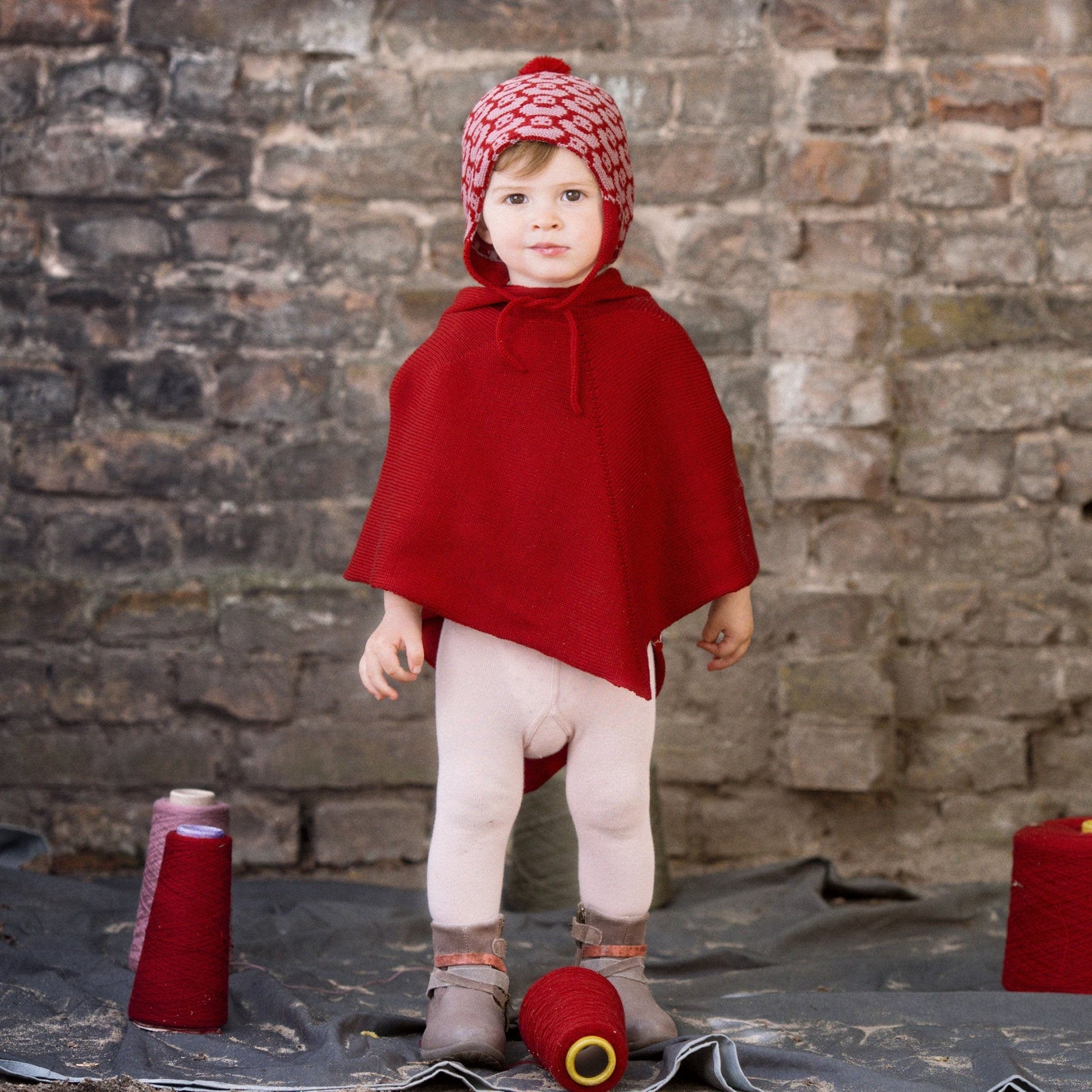 Ein kleines Mädchen trägt über ihrer Strumpfhos ein gestricktes rotes Cape mit Kapute. Auf dem Kopf hat es eine rote Mütze mit rosa Entschenmuster