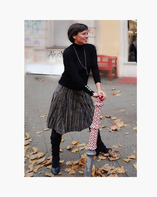 Eine Schwarzhaarige Frau mit schwarzem Pullover stützt sich auf die bunt bestrickten Straßen-Pöller. Sie trägt einen Melierten Rock in Schwarz/Beige