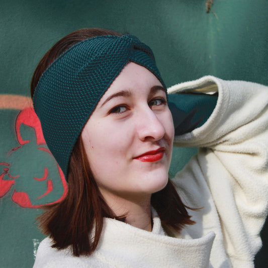 Eine Junge Frau trägt ein Petrol Farbenes Stirnband mit einer Turban-ähnlichen Verdrehung. Gestrickt im  Doubleface Patentmuster, außen Knötchenstruktur und Innen sieht es wie ein klassisch rechts gestricktes Muster aus.